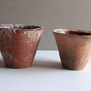 Peut inclure: Deux pots de fleurs en terre cuite patinée de forme conique. Le plus grand pot à gauche présente une surface rugueuse et texturée avec des dépôts minéraux blancs. Le plus petit pot à droite a une finition brun-rouge plus lisse et un bord irrégulier.