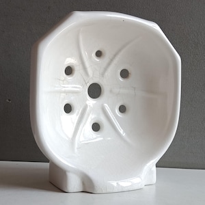 May include: A white ceramic soap dish with a shell-like shape, featuring a central drainage hole surrounded by six smaller holes arranged in a radial pattern. The surface has a glossy finish with visible fine crackle glaze detailing throughout.