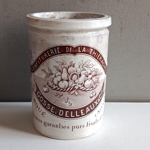 Puede incluir: Un tarro de cerámica cilíndrico vintage con un aspecto ligeramente desgastado. El tarro es principalmente blanquecino con una etiqueta granate descolorida con texto y una ilustración de una cesta de frutas. El texto incluye "CONFITURE DE LA THIERACHE" y "GOSSE. DELLEAUX&C".
