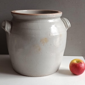 Peut inclure: Un grand pot en céramique gris clair doté de deux anses latérales et d'une large ouverture. L'extérieur présente de subtiles stries horizontales près du bord et une légère décoloration jaunâtre. Une pomme rouge et jaune est posée à côté.
