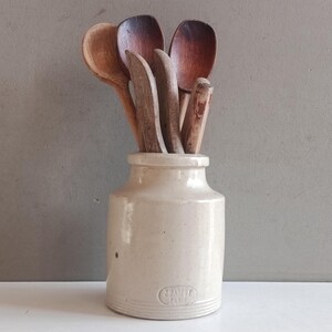 May include: A cream-colored ceramic crock with a cylindrical body and a small rim, featuring the embossed text LEA & PERRINS. Inside the crock are several wooden kitchen utensils, including three rounded spoons and two flat-edged spatulas of varying brown tones.