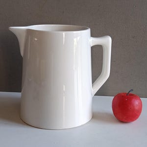 Pode incluir: Um jarro de cerâmica branco com uma alça e bico, ao lado de uma maçã vermelha. O jarro tem um acabamento liso e brilhante. A maçã é de uma cor vermelha vibrante. O fundo é cinza neutro.