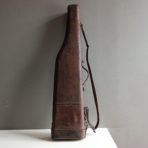Könnte beinhalten: Ein Vintage-Gewehrkoffer aus braunem Leder mit Schulterriemen. Der Koffer hat eine rechteckige Basis und verjüngt sich nach oben, mit einem Griff und Schnallen Detail. Das Leder hat ein abgenutztes, strukturiertes Aussehen, das auf Alter und Gebrauch hindeutet. Der Koffer dient zum Schutz und Transport einer Schusswaffe.
