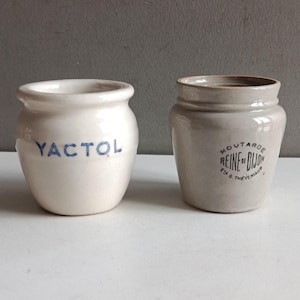 以下が含まれることがあります： 2つのアンティークセラミックジャー。1つは白で、青い文字で「YACTOL」と書かれています。もう1つはベージュで、黒い文字で「MOUTARDE REINE DE DIJON」と書かれています。どちらのジャーもヴィンテージ感があります。