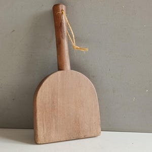 Può includere: Una paletta di legno con un manico e una testa curva a forma di pala. La paletta è realizzata in legno marrone chiaro e ha un piccolo foro nella parte superiore del manico con un pezzo di spago attaccato. La paletta viene probabilmente utilizzata per scopi culinari o di cottura.