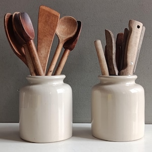 Pode incluir: Dois potes de cerâmica em tom off-white contendo vários utensílios de cozinha. O pote da esquerda contém várias colheres de madeira castanho-escuras e uma espátula plana. O pote da direita contém facas com cabos de madeira clara e ferramentas.