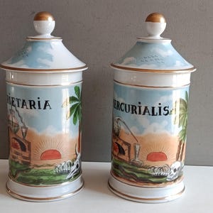 May include: Two antique ceramic apothecary jars with lids. The jars feature a light blue and white design with gold accents. Each jar has a unique illustration with text, a sunset, and a palm tree.