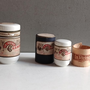 May include: Two white ceramic jars with lids, one dark blue cylindrical container, and a rolled paper label, all featuring vintage-style branding with the text La Reine des Cremes and La Reine, including portraits and decorative red and black typography.