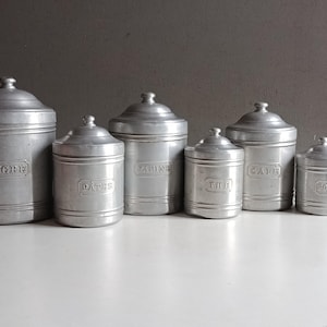 Peut inclure: Ensemble de six bocaux métalliques vintage avec couvercles, disposés sur une surface blanche. Les bocaux sont étiquetés avec les mots français : "SUCRE", "DATES", "FARINE", "THE", "CAFE" et "POIVRE". Les bocaux ont une finition argentée.