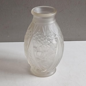 Puede incluir: Un pequeño jarrón de vidrio esmerilado con cuerpo redondeado y cuello estrecho. El jarrón presenta un diseño en relieve detallado de hojas y bayas. El vidrio transparente permite el paso de la luz, resaltando los intrincados patrones. El jarrón mide unos 10 cm de alto.