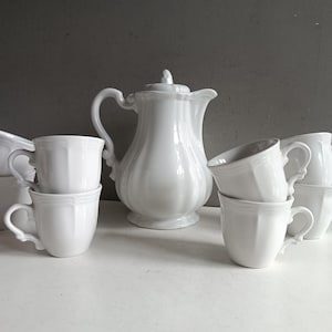 May include: A set of white porcelain teacups and a teapot. The teapot has a fluted design and a lid with a knob. The teacups have handles and are stacked in pairs.