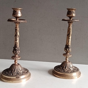 Könnte beinhalten: Zwei antike Messingkerzenständer mit aufwendigen Blumen- und Blattdetails. Jeder Kerzenständer hat einen breiten, dekorativen Sockel, einen schlanken Stiel mit geschnitzten Akzenten und eine becherförmige Oberseite zur Aufnahme einer Kerze. Die Kerzenständer stehen auf einer weißen Oberfläche.