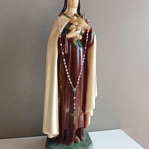 May include: A religious statue of a nun wearing a dark brown habit and a cream-colored cloak. The figure holds a bouquet of flowers and a crucifix in the hands, with a pink beaded rosary draped around the neck and hanging down the front.