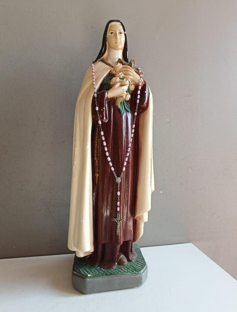 May include: A religious statue of a nun wearing a dark brown habit and a cream-colored cloak. The figure holds a bouquet of flowers and a crucifix in the hands, with a pink beaded rosary draped around the neck and hanging down the front.