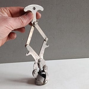 Könnte beinhalten: Ein silberfarbener Korkenzieher im Vintage-Stil mit einem T-förmigen Griff und einem scharnierartigen, rautenförmigen Mechanismus. Der Korkenzieher hat einen Flaschenöffner und eine Schraube zum Entfernen von Korken. Das Wort "WONDERTOOL" ist auf dem Werkzeug eingeprägt.