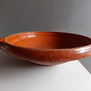 May include: A rustic, shallow, terracotta-colored ceramic bowl with a small handle. The bowl has a wide, rounded shape and a glossy finish. The interior and exterior are the same color. The bowl is likely used for serving food.