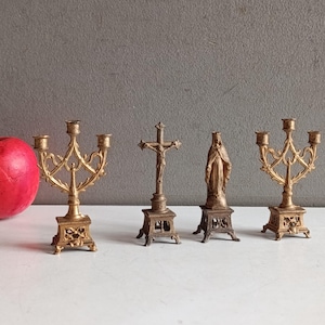 Puede incluir: Naturaleza muerta con figuras religiosas y candelabros. La colección incluye dos candelabros dorados ornamentados, un crucifijo y una estatua de la Virgen María. A la izquierda hay una gran manzana roja.