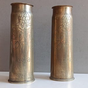 Puede incluir: Dos casquillos de proyectil cilíndricos de latón colocados en posición vertical, cada uno con un diseño floral grabado de una flor similar a un cardo con hojas y tallos. El metal tiene una pátina marrón dorada con iniciales grabadas en la base.