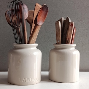 May include: Two off-white ceramic crocks holding kitchen utensils. The left crock features the embossed text LAB Lagny and contains a wire whisk and wooden spoons. The right crock holds several wooden-handled knives and flatware pieces.