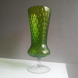 May include: A tall, green glass vase with a textured surface and a flared top. The vase has a bulbous base and a clear, circular foot. The overall height is approximately 25 cm.
