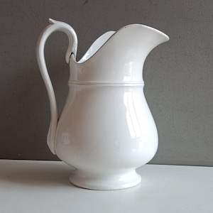 Può includere: Una brocca in ceramica bianca con finitura liscia e lucida, manico curvo, ampio beccuccio e corpo arrotondato che si restringe verso una base circolare. Il design è semplice e funzionale, caratterizzato da un colore bianco uniforme.