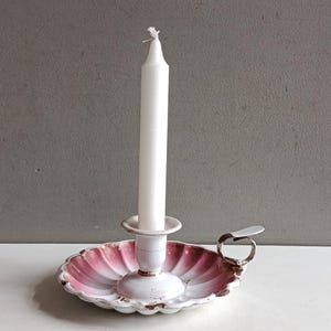 May include: A white candle in a vintage candle holder. The holder features a scalloped, pink and white dish with a handle. The candle holder is metal and has a distressed, antique appearance. The candle is white.