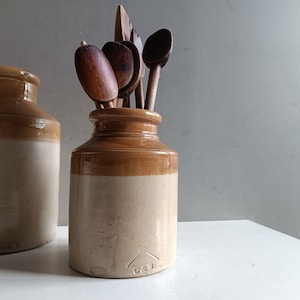 Op de afbeelding: Twee bruine steengoed potten met een witte glazuur. De kleinere pot heeft houten lepels en spatels erin.
