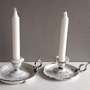 Può includere: Due candelabri in smalto bianco con disegni floreali e manici. Ogni candelabro ha una candela bianca. I candelabri sono su una superficie bianca con sfondo grigio. I candelabri hanno un aspetto vintage e usurato.