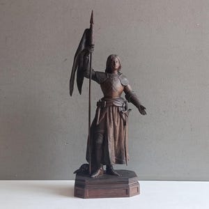 Könnte beinhalten: Eine dunkelbraune Metallstatue einer Figur in mittelalterlicher Rüstung auf einem sechseckigen Sockel. Die Figur hält einen langen Speer mit einer drapierten Flagge in der einen Hand und streckt die andere Hand aus. Die Rüstung umfasst einen Brustpanzer.