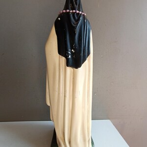 May include: A standing figurine of a figure wearing a long cream-colored robe and a black head covering. A string of small pink beads rests around the neck area. The figure is positioned on a rectangular base with a textured green surface.