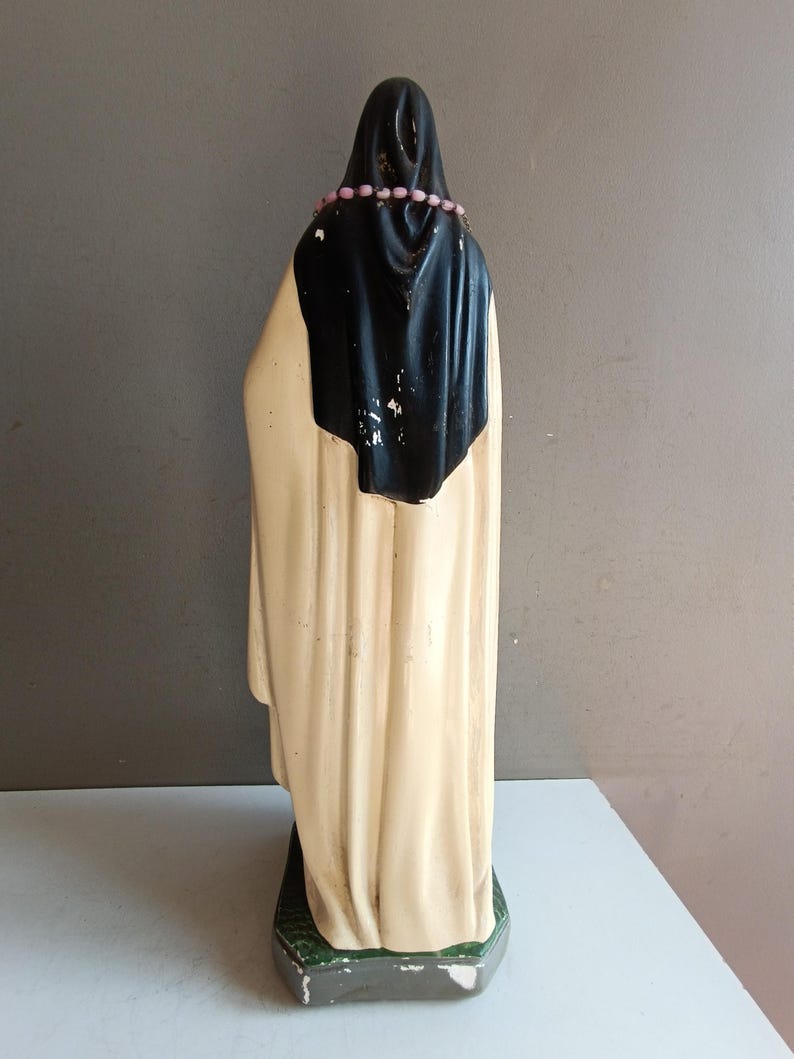 May include: A standing figurine of a figure wearing a long cream-colored robe and a black head covering. A string of small pink beads rests around the neck area. The figure is positioned on a rectangular base with a textured green surface.