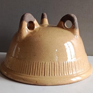 Op de afbeelding: Een beige keramische cloche met een afgeronde vorm en verticale lijnen aan de basis. Het heeft twee gebogen handvatten met cirkelvormige openingen en een centrale knop. De handvatten en knop zijn donkerbruin.