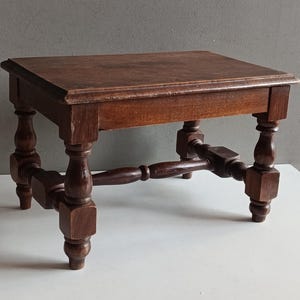 May include: A small rectangular wooden stool with a dark brown finish. The stool features four legs with turned detailing and square block joints, connected by a central horizontal stretcher bar that provides structural support to the base.
