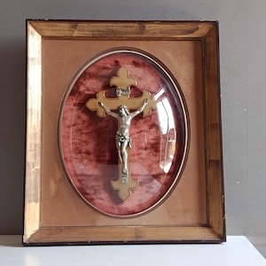 May include: A framed wall crucifix featuring a silver-toned metal figure of Jesus mounted on a light-colored wooden cross. The cross is set against a deep red velvet background within an oval glass display case, enclosed in a gold-toned frame.
