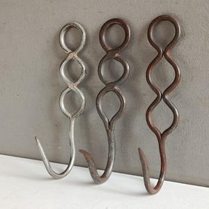 Op de afbeelding: Drie metalen haken met een uniek, decoratief ontwerp. De haken zijn gemaakt van ijzer en hebben een gedraaid, golvend patroon. De haken zijn in verschillende tinten zilver, bruin en roest, en zijn geschikt voor het ophangen van items.