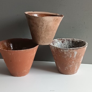 May include: Three terracotta pots of varying sizes and shapes. The pots are all unglazed and have a rustic, earthy appearance. The largest pot is the most weathered, with a white, chalky residue on its surface.