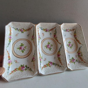 Peut inclure: Un plat de service divisé en porcelaine blanche avec un motif floral. Le plat comporte trois sections, chacune avec une rose rose dans un cercle doré, entourée de fleurs colorées et de feuilles vertes. Les bords sont dorés.