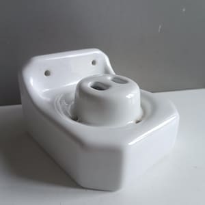 Op de afbeelding: Witte, wandgemonteerde tandenborstelhouder van keramiek met twee gaten voor tandenborstels. De houder heeft een afgerond, licht afgeschuind ontwerp.