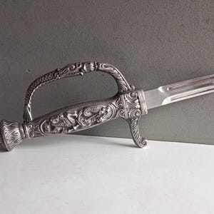 May include: Ornate silver letter opener with a curved handle and detailed carvings. The blade is straight and rectangular. The handle features a decorative design with floral and mythical creature motifs. The letter opener is displayed on a white surface.