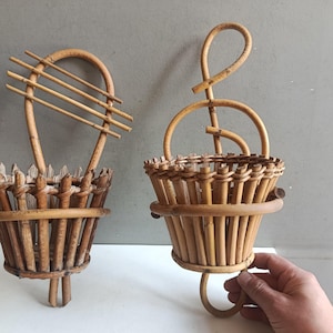 Peut inclure: Deux paniers muraux vintage en rotin tressé. L'un a une poignée incurvée avec des lattes horizontales, l'autre un motif de clé de sol. Les deux ont une forme de panier conique, parfait pour exposer des plantes ou des objets décoratifs.
