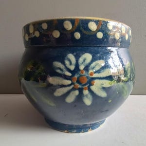 Könnte beinhalten: Ein blauer Keramiktopf mit floralem Design. Der Topf hat eine große weiße Blume mit orangefarbenem Zentrum auf der Vorderseite. Der Rand ist mit weißen Punkten und orangefarbenen Akzenten verziert. Der Topf hat eine abgerundete Form und einen leicht ausgestellten Boden.