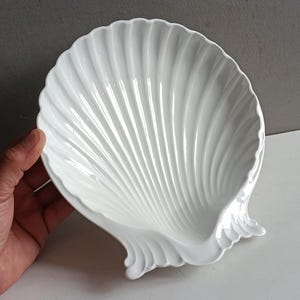 May include: A white ceramic dish shaped like a large scallop shell. The bowl features a glossy finish with deep, radiating fluted ridges that extend from the base to the scalloped edge, creating a textured, fan-like appearance for this serving piece.