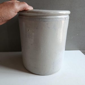 Op de afbeelding: Een cilindervormige, lichtgrijze stenen pot met een gladde, geglazuurde afwerking en een decoratieve kralenstructuur langs de bovenrand. Het vat heeft een stevige, dikwandige constructie met een eenvoudig en functioneel ontwerp.