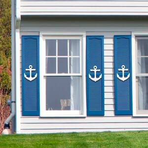 May include: A white house with three windows with blue shutters and white anchor accents. The windows are on the front of the house and have white trim. The house is surrounded by green grass.