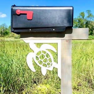 May include: A black mailbox with a red flag is mounted on a wooden post with a white sea turtle cutout. The mailbox is in front of a grassy area with a body of water in the background.