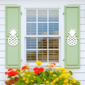 May include: A white house with green shutters featuring pineapple cutouts. The windows have white trim and are surrounded by a planter box filled with yellow and red flowers.