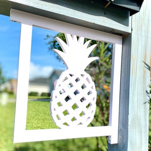 May include: White wooden pineapple cutout mounted on a wooden post. The pineapple is a decorative element for a mailbox or other outdoor structure.