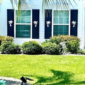 May include: A white house with blue shutters and palm tree cutouts. The house has a green lawn and bushes in front of it.