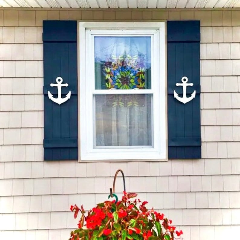 May include: A white window with blue shutters and white anchor accents. The window has a stained glass design in the center. A red flower pot is in front of the window.