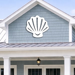 May include: A white seashell-shaped wall decoration mounted on a blue-gray shingled wall. The shell is a simple, stylized design with radiating lines and a curved base, suggesting a coastal or nautical theme. The house has white trim and a black lantern.